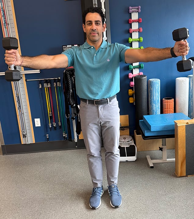 Man standing holding weights with his arms raised upward about 90 degrees and apart from each other about 45 degrees.