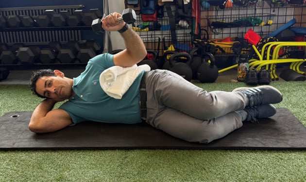 Man lying on his right side, with his right hand cradling his head and his left hand  holding a weight raised upward. A towel is nested between his left elbow and his body.
