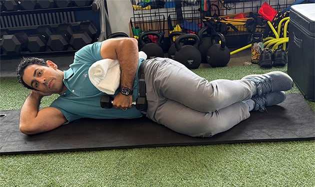 Man lying on his right side, with his right hand cradling his head and his left hand  holding a weight. A towel is nested between his left elbow and his body.