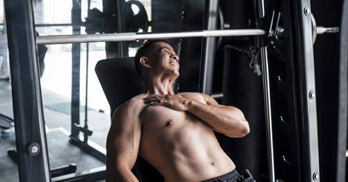 A man on a weight bench grimacing and holding his pectoral muscle.